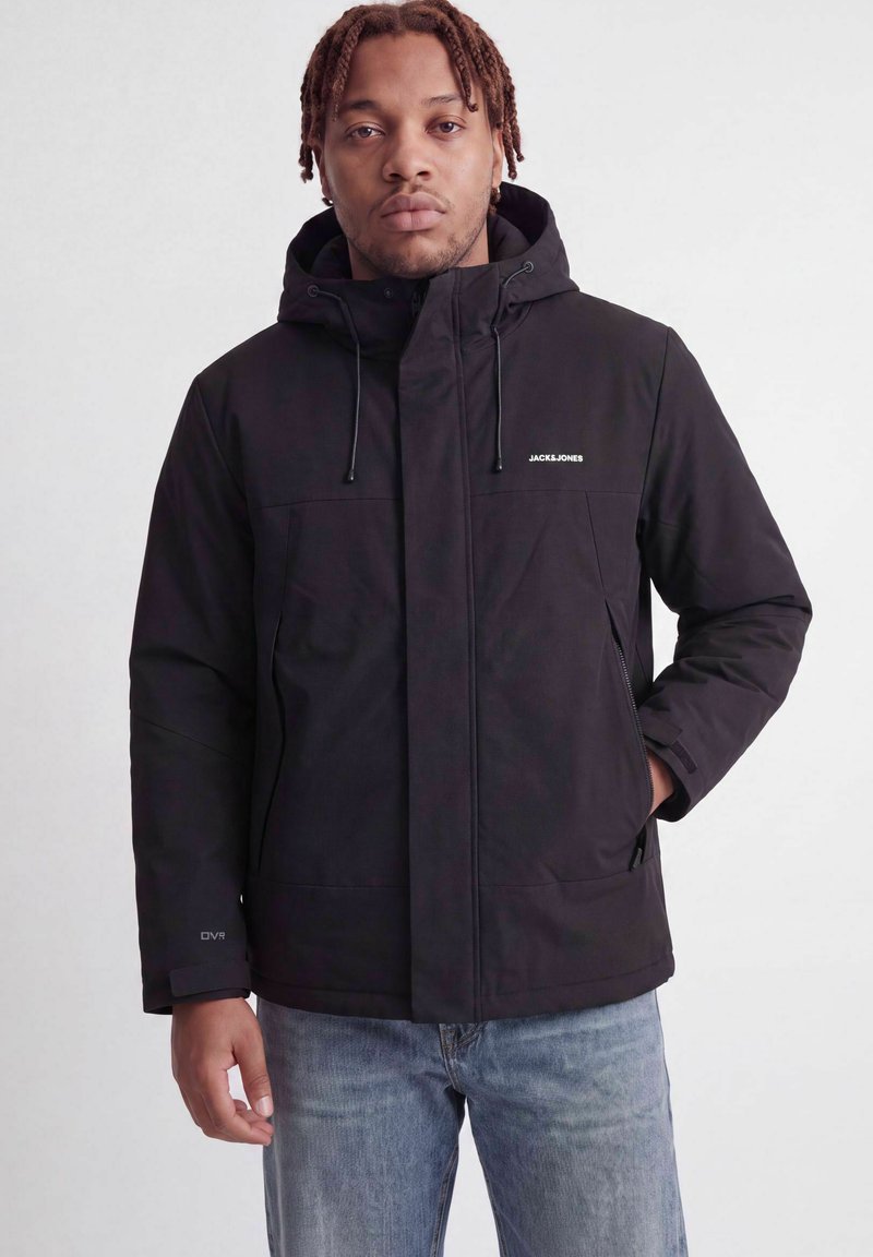Homme aux cheveux tressés, portant une veste noire à capuche Jack & Jones et un jean bleu, debout devant un fond clair uni.