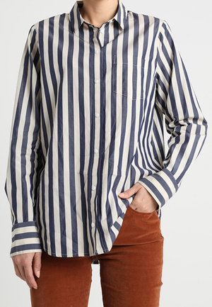 Person trägt ein langärmliges, blau-weiß vertikal gestreiftes Hemd mit einer Hand in der Tasche einer rostfarbenen Hose.