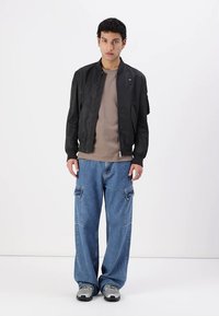Giubbotto bomber nero con una finitura liscia, t-shirt beige sotto, e pantaloni cargo in denim blu a taglio largo con tasche frontali. Sneakers grigie sportive.
