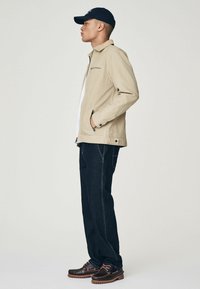 Beige jack met een rits, zijzakken en een kraag; donkerblauwe jeans met helderdere stiksels; bruine schoenen met opvallende veters en een gestructureerde zool.