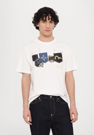T-shirt en coton blanc avec un design graphique affichant quatre poches colorées en bleu, noir et beige, ainsi que divers détails de couture.