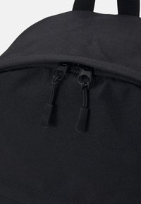 Gros plan sur un sac à dos en tissu noir montrant deux tirettes de fermeture éclair sur le compartiment principal avec des languettes en tissu et une sangle de poignée supérieure.