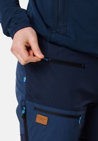 Marina cargo byxor med en ficka med dragkedja, accentuerad av en blå dragkedja och en brun läderetikett. Svensk flagga emblem på sidan.