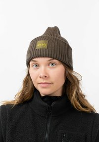 Bruine geribbelde gebreide beanie met een puntige bovenkant en een geel logo-opdruk. Het model draagt een zwart fleece vest en toont een natuurlijke huidtoon.