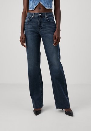 Vrouwelijk model dat donkerblauwe, rechtgesneden jeans met hoge taille en rafelige zomen draagt, zwarte puntige hakken, en een cropped blauwe denim vest.