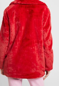 Rød falsk pelsjakke med en blød, plysset tekstur, der har en bred krave og en løs pasform. Synlige sømme og ingen knapper.