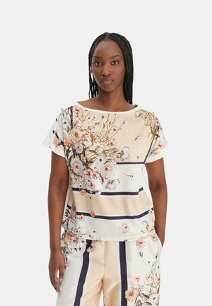 Donna che indossa una maglia color crema e pantaloni con motivi floreali e a righe, in piedi con le mani in tasca contro uno sfondo chiaro e uniforme.