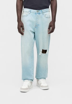 Mężczyzna w jasnoniebieskich dżinsach typu szerokie nogawki z dużą podartą dziurą na prawym kolanie, białe trampki i czarną koszulę.