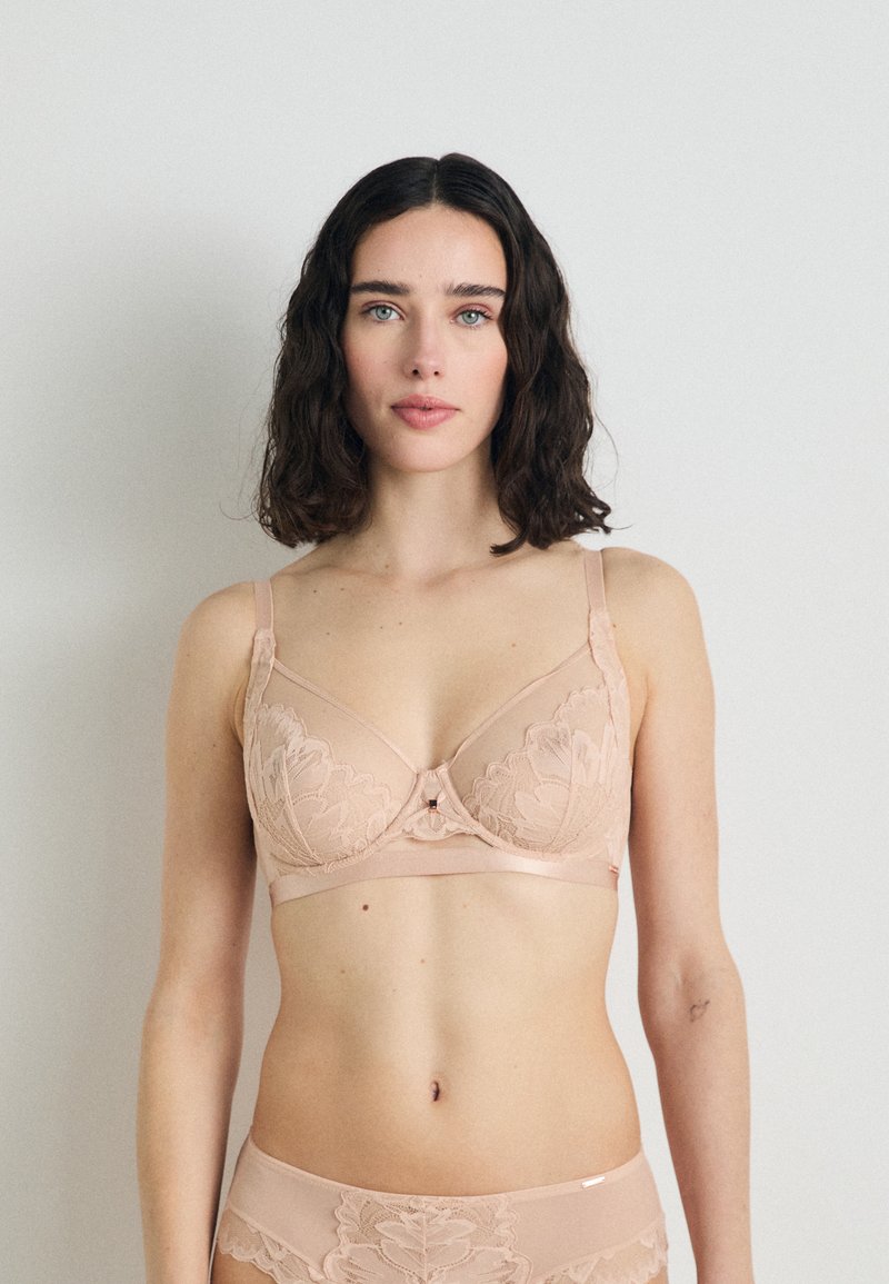 Bielizeň v mäkkom nude materiáli s kvetinovými čipkovými dizajnmi, s kosticami na podporu, nastaviteľnými ramienkami a zapínaním vpredu. Zodpovedajúce nohavičky s kvetinovými akcentmi.