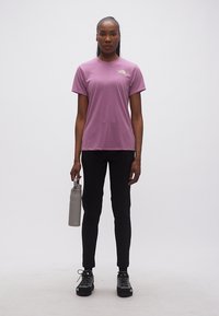 Γυναίκα όρθια, φοράει μοβ μπλούζα με κοντά μανίκια, μαύρο παντελόνι, μαύρα παπούτσια, κρατώντας ένα γκρι παγούρι νερού στο πλάι της.