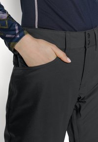 Personne portant un pantalon noir avec la main partiellement dans la poche avant et une chemise bleue à motifs à manches longues visible sur le bras.