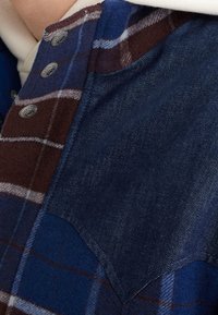 Plaid shirt featuring blue and brown tones with denim accents. Soft texture, metal buttons, and a distinct pointed yoke design.