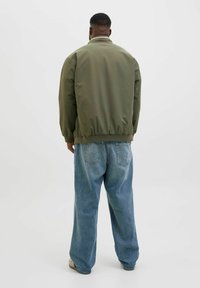 Zelená bomber bunda s žebrovaným límcem a manžetami, v kombinaci s volně střiženými světle modrými džíny. Bunda má hladkou, lesklou texturu.