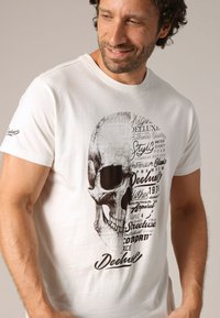 T-shirt en coton blanc avec un graphique de crâne noir et divers textes dans un mélange de polices. Manches courtes et col rond.