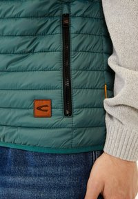 Gepolstertes grünes Westchen mit einer Reißverschluss-Tasche, braunem Leder-Logo-Patch, verstellbarem Saum und darunter einem strukturierten grauen Pullover.