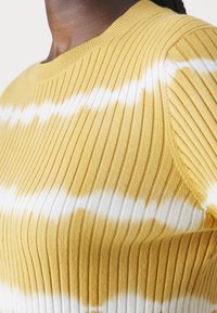 Żółty sweter dzianinowy z prążkowaną fakturą, z białymi wzorami tie-dye, krótkimi rękawami i okrągłym dekoltem. Miękka tekstura z widocznymi prążkami.