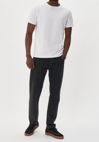 Mann trägt ein schlichtes weißes T-Shirt mit kurzen Ärmeln, ausgewaschene schwarze Jeans und schwarze Sneaker mit braunen Sohlen, steht mit einer Hand in der Tasche.