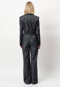 Vrouw met steil blond haar in een donkergrijze maatpak met een taillerende blazer en brede broek, staande met de rug naar de kijker gekeerd.