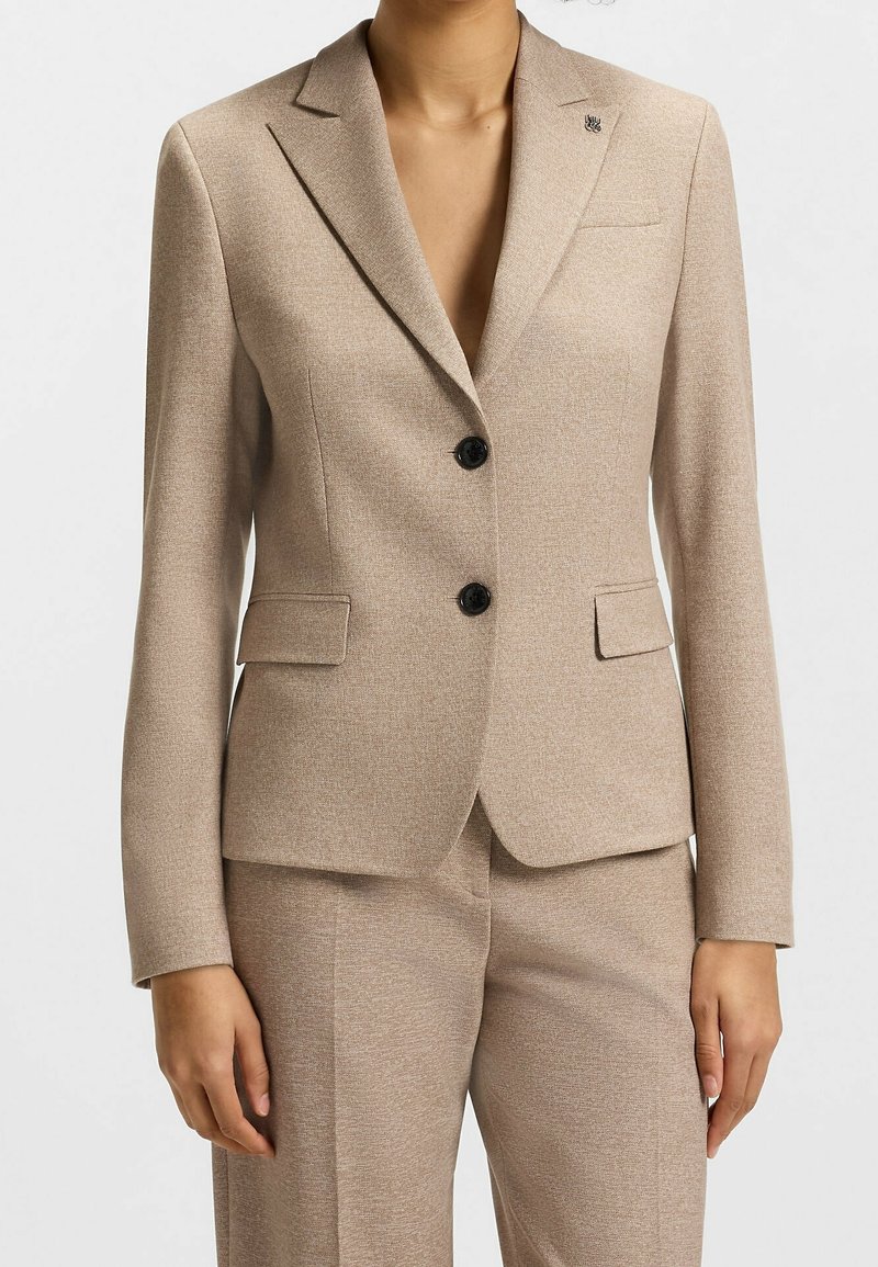 Femme portant un blazer beige à deux boutons avec revers crantés et un pantalon assorti, vue du cou jusqu'à mi-cuisse sur un fond uni.