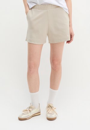 Άτομο που φοράει μπεζ σορτς με ελαστική μέση, λευκές κάλτσες crew και μπεζ αθλητικά παπούτσια σε απλό λευκό φόντο.