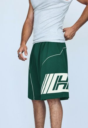 Homme portant un débardeur gris et un short de basket vert avec des rayures blanches et de grandes lettres blanches sur le côté.