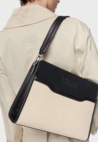 Płócienna torba na ramię o kremowym korpusie i czarnym wierzchu, z wytłoczonym logo "TOUS", skórzanymi akcentami oraz regulowanym paskiem.