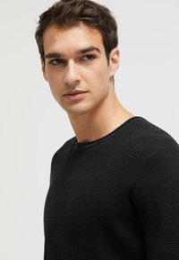 Black textured sweater with a round neck and ribbed detailing, displayed on a model against a neutral background.
