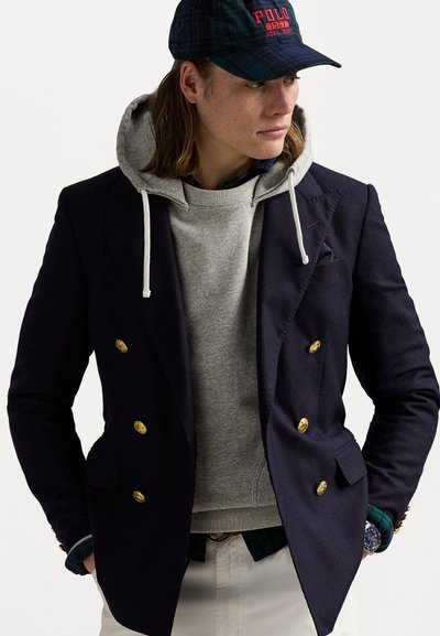 Blazer croisé bleu marine avec des boutons dorés superposé sur un sweat à capuche gris. Des cheveux longs et une casquette à carreaux complètent le look.