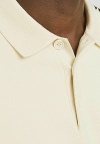 Krämfärgad polo skjorta med ribbad krage och enstaka knäppning. Har texturerat tyg och detalj längs sidan.
