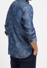 Denim shirt in donkerblauw met een lichter bloemenpatroon, lange mouwen opgerold en een gebogen zoom. De stof lijkt glad en zacht.