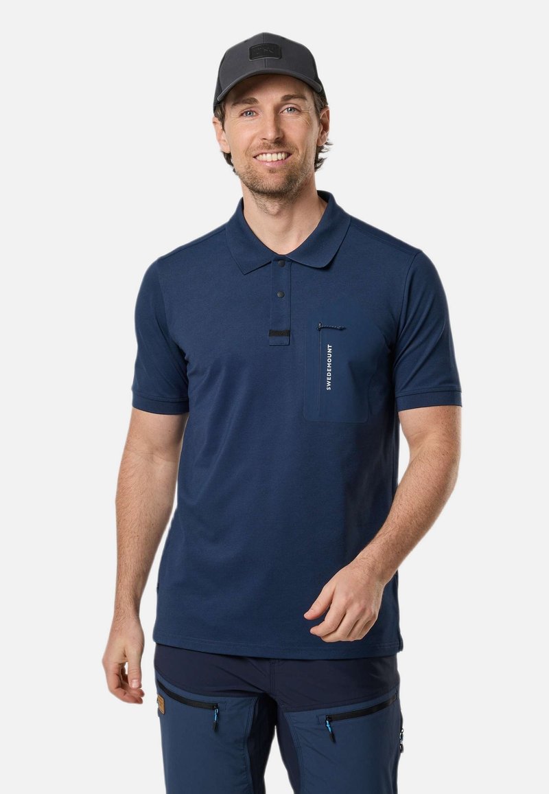Homme portant un polo bleu marine avec une poche poitrine zippée et une casquette sombre, souriant sur fond blanc uni.