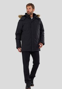Man in een zwarte winterparka met een bontgevoerd capuchon, zwarte jeans en zwarte laarzen, staande tegen een effen lichtgrijze achtergrond.