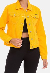 Yellow denim jacket with a fitted design, silver hardware, two chest pockets, and stitching details. Worn over a black top.