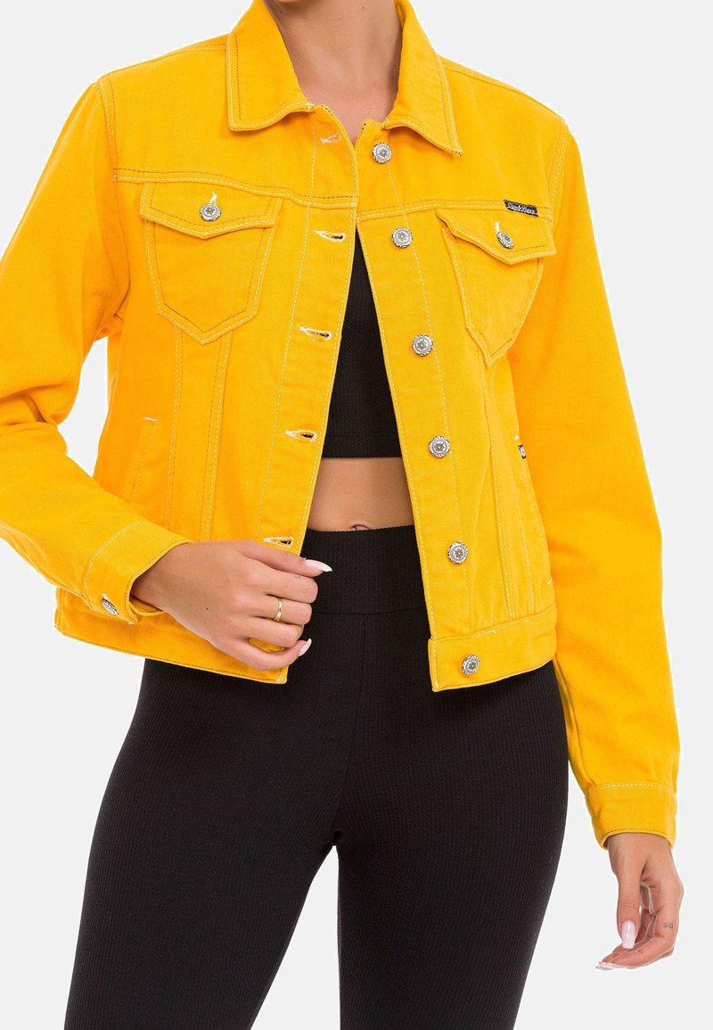Yellow denim jacket with a fitted design, silver hardware, two chest pockets, and stitching details. Worn over a black top.