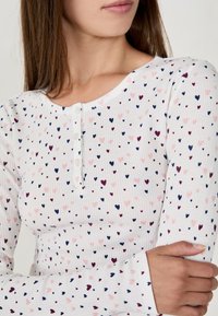 Kobieta w białej bluzce z długim rękawem, pokrytej małymi różowymi, granatowymi i bordowymi wzorami w serca oraz trzema białymi guzikami w pobliżu dekoltu.