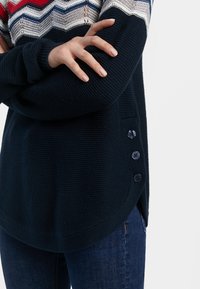 Marineblauer Strickpullover mit einem gestreiften, bunten Muster am Kragen. Verfügt über seitliche Schlitze und vier dunkle Knöpfe auf der linken Seite.