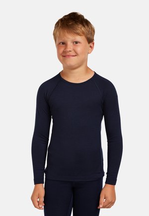 Jonge jongen met lichtbruin haar, gekleed in een nauwsluitend marineblauw shirt met lange mouwen en bijpassende broek, staand tegen een effen achtergrond.