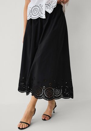 Kobieta w czarnej spódnicy midi z falistym kwiatowym wycięciem na dole i czarnych sandałach na obcasie z paskami na jednolitym tle.