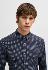 Granatowa koszula z trójkątnym białym wzorem, z długim rękawem, z kołnierzykiem zapinanym na guziki oraz białymi guzikami. Wykonana z lekkiego materiału.