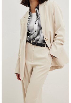 Kobieta ubrana w beżowy garnitur z wysokim stanem spodni, czarnym paskiem, białym topem na ramiączkach oraz czarno-białą koszulą w pionowe paski na guziki.