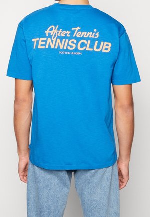 Person wearing a blue t-shirt with "After Tennis TENNIS CLUB SCOTCH & SODA" text printed on the back, paired with light denim jeans.