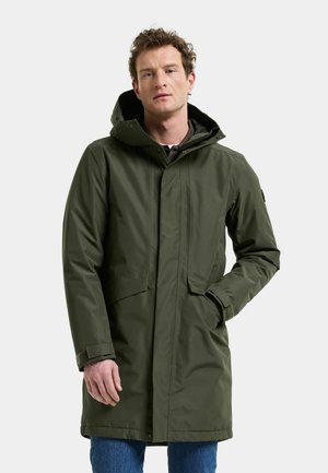 Mand iført en lang olivengrøn hættetrøje med forlommer og blå jeans mod en ensartet lys baggrund.