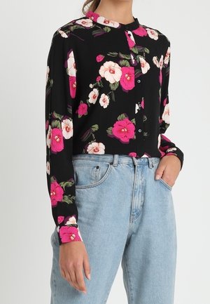 Vrouw die een zwart, knoopsluitend shirt met lange mouwen draagt met een roze-witte bloemenprint, gecombineerd met lichtblauwe, hooggetailleerde spijkerbroek.
