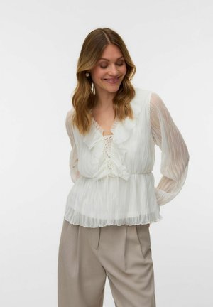 Kobieta o falowanych włosach ubrana w przezroczystą białą bluzkę z falbankami i beżowe plisowane spodnie, uśmiechająca się i patrząca w dół z rękami za plecami.