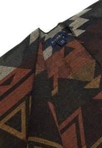 Wollschal mit geometrischem Muster in Dunkelgrau, Rost und Creme. Die Textur wirkt weich, mit einem umgeschlagenen Rand, der das Etikett zeigt.