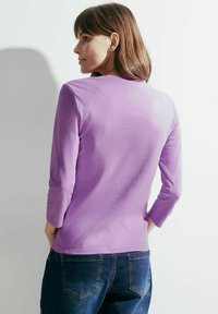 Jasnofioletowa bluzka z długim rękawem, okrągłym dekoltem i rękawami do trzech kwart, w połączeniu z ciemnoniebieskimi dżinsami. Gładki, elastyczny materiał.