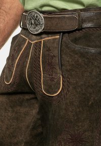 Pantalones cortos de cuero marrón con intrincados bordados en burdeos, un diseño trenzado y una hebilla de cinturón decorativa plateada con el año "1880."