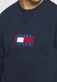 Marinblå sweatshirt i bomull, med en rektangulär broderad logotyp i rött, vitt och blått på bröstet.