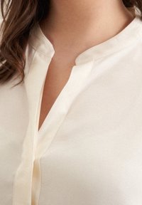 Krémová blůza s mandarínkovým límečkem a saténovou texturou, s předním zavíráním a vertikálními švy.