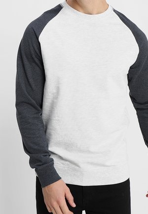 Hombre vestido con una camisa blanca de manga larga con mangas raglán gris oscuro y pantalones negros, de pie frente a un fondo claro.